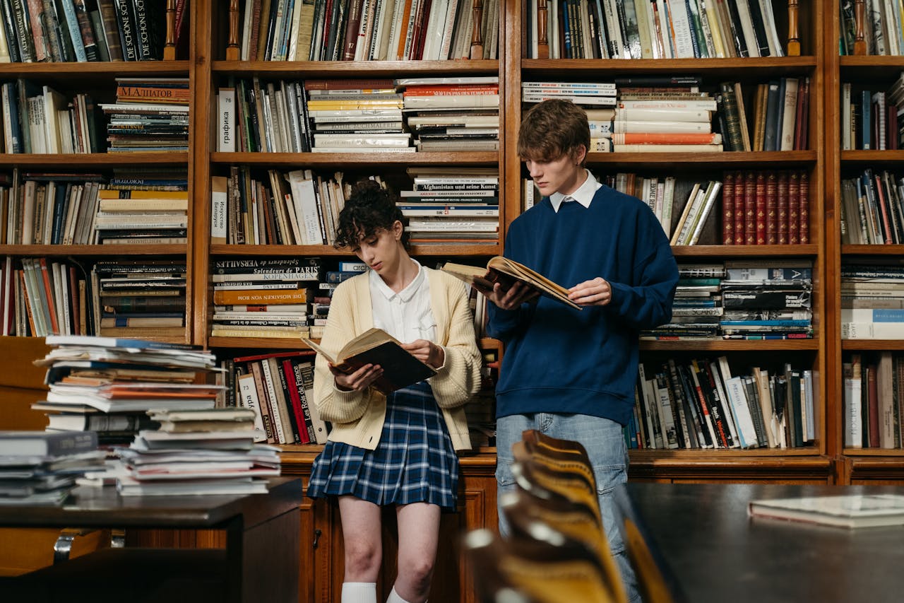 Two students reading books in a library filled with shelves of stacked books