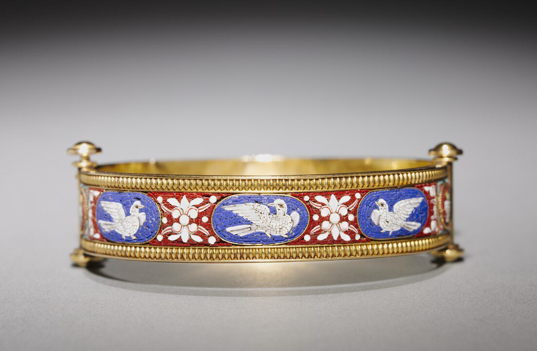 Gold bracelet with a rectangular enamel panel design, featuring alternating white dove and floral motifs, blue and red background, raised gold edges with spherical accents on each corner, displayed against a neutral gradient background