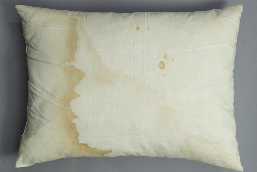 Stained pillow, white fabric with visible yellow and brown discoloration, standard rectangular shape, soft texture, resting on bed or flat surface, signs of wear and aging