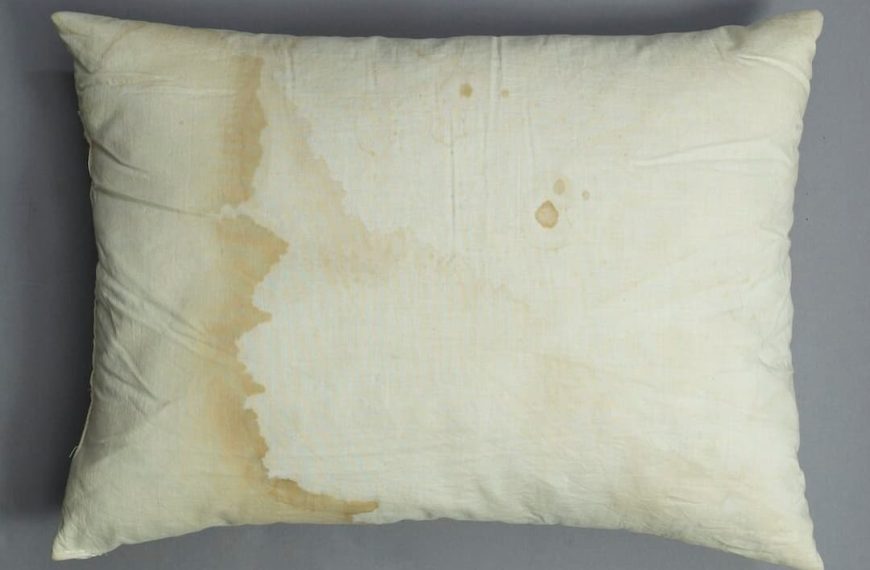 Stained pillow, white fabric with visible yellow and brown discoloration, standard rectangular shape, soft texture, resting on bed or flat surface, signs of wear and aging