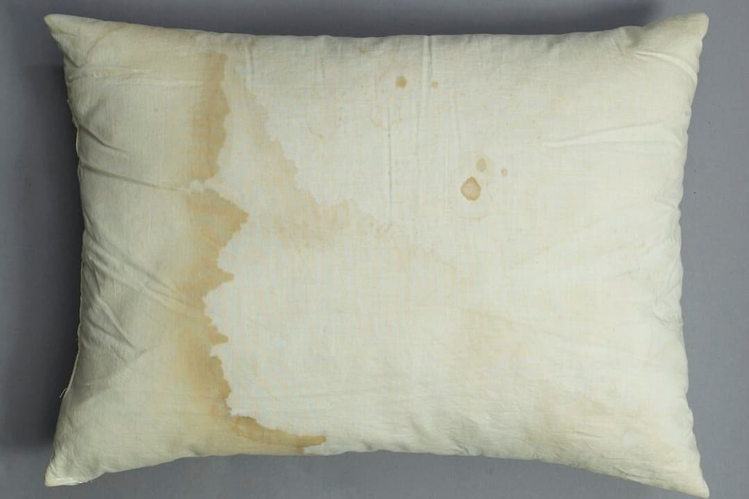 Stained pillow, white fabric with visible yellow and brown discoloration, standard rectangular shape, soft texture, resting on bed or flat surface, signs of wear and aging