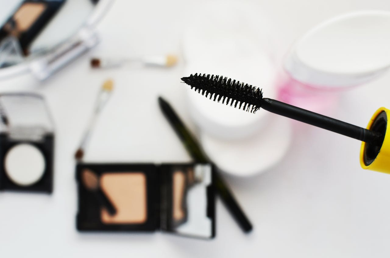Close-up of a black mascara wand with various makeup items blurred in the background on a white surface