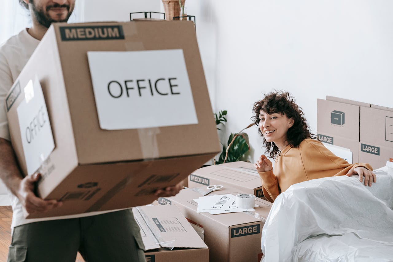 Smiling woman sitting on covered furniture, man holding box labeled "Office", surrounded by moving boxes, indoors