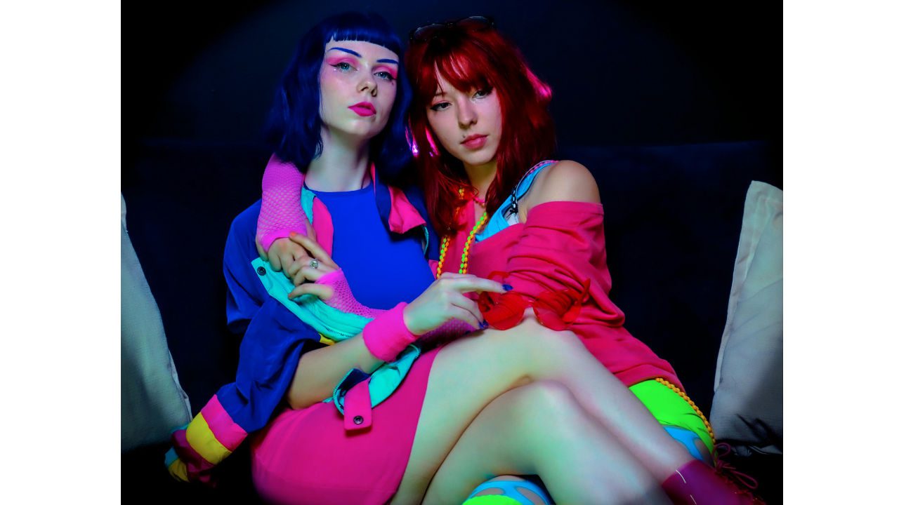 Two women in vibrant neon clothing, bold makeup, sitting together on a couch in a dark room