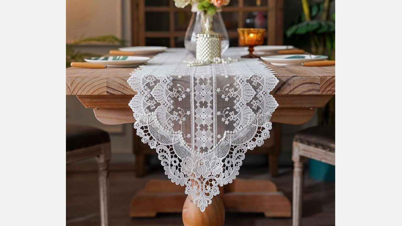 wooden dining table, white lace table runner, floral patterns, plates with gold cutlery, centerpiece vase with flowers, candle decor, cozy indoor setting