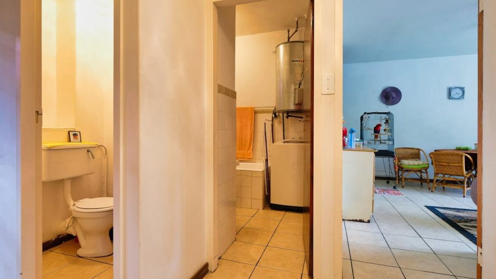 Interior view of a modest home, showing a bathroom with a toilet and sink on the left, a narrow hallway with a washing machine in the center, and a living room on the right with retro-style furniture, wall art, and a coffee table, tiled floors throughout, warm and cool lighting contrast