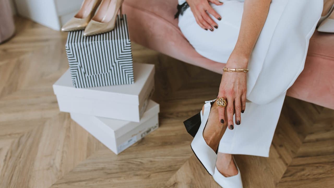 Woman sitting on a pink chair, wearing white pants and white heels, holding her ankle in discomfort, shoe boxes and a handbag nearby, wooden herringbone floor, indoor setting suggests shoe shopping or foot pain