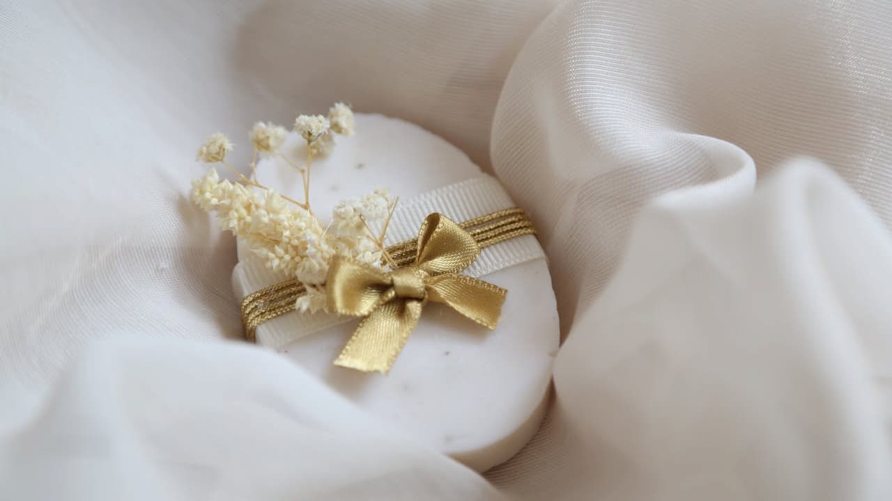 white oval soap bar, wrapped with gold ribbon, small dried flowers, elegant fabric background, soft lighting, gift-like presentation