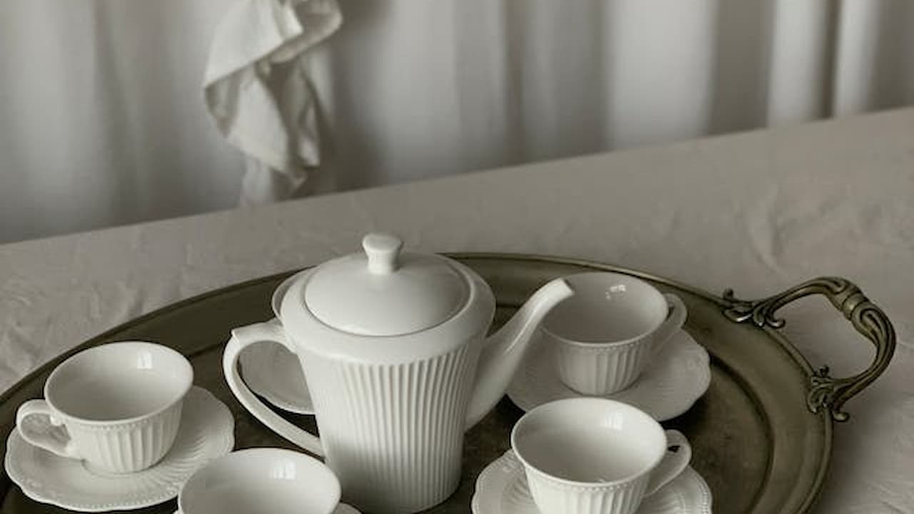 White porcelain teapot with matching teacups and saucers arranged on a round metallic tray, set on a light-colored tablecloth, soft curtain backdrop, minimal and elegant tea service setup