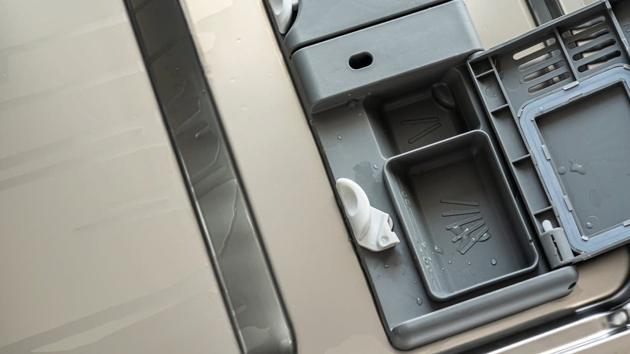 Open dishwasher detergent compartment, gray plastic dispenser, some detergent residue visible, stainless steel interior background