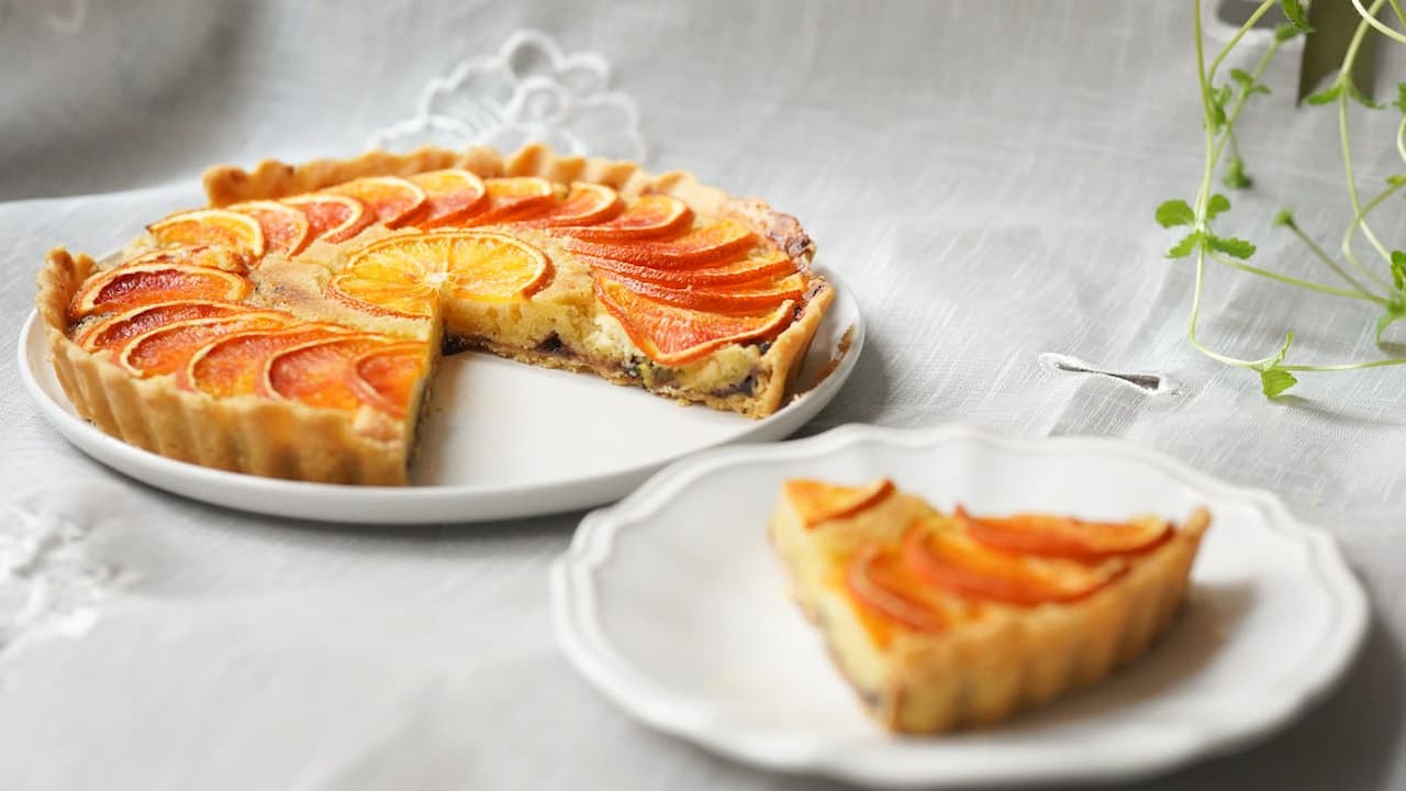 Peach tart on a white plate, one slice served separately, golden crust, neatly arranged peach slices on top, set on a white tablecloth, small green plant partially visible in the corner.