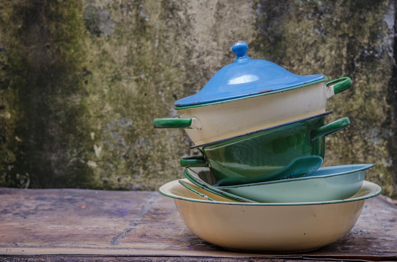 Stack of old enamel cooking pots, blue lid on top, green and cream pans, resting on wooden surface, mossy wall background