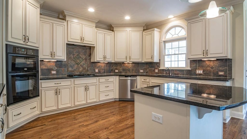 Spacious modern kitchen with white cabinets, black granite countertops, stainless steel appliances, tile backsplash, center island with built-in sink, hardwood flooring, arched window letting in natural light