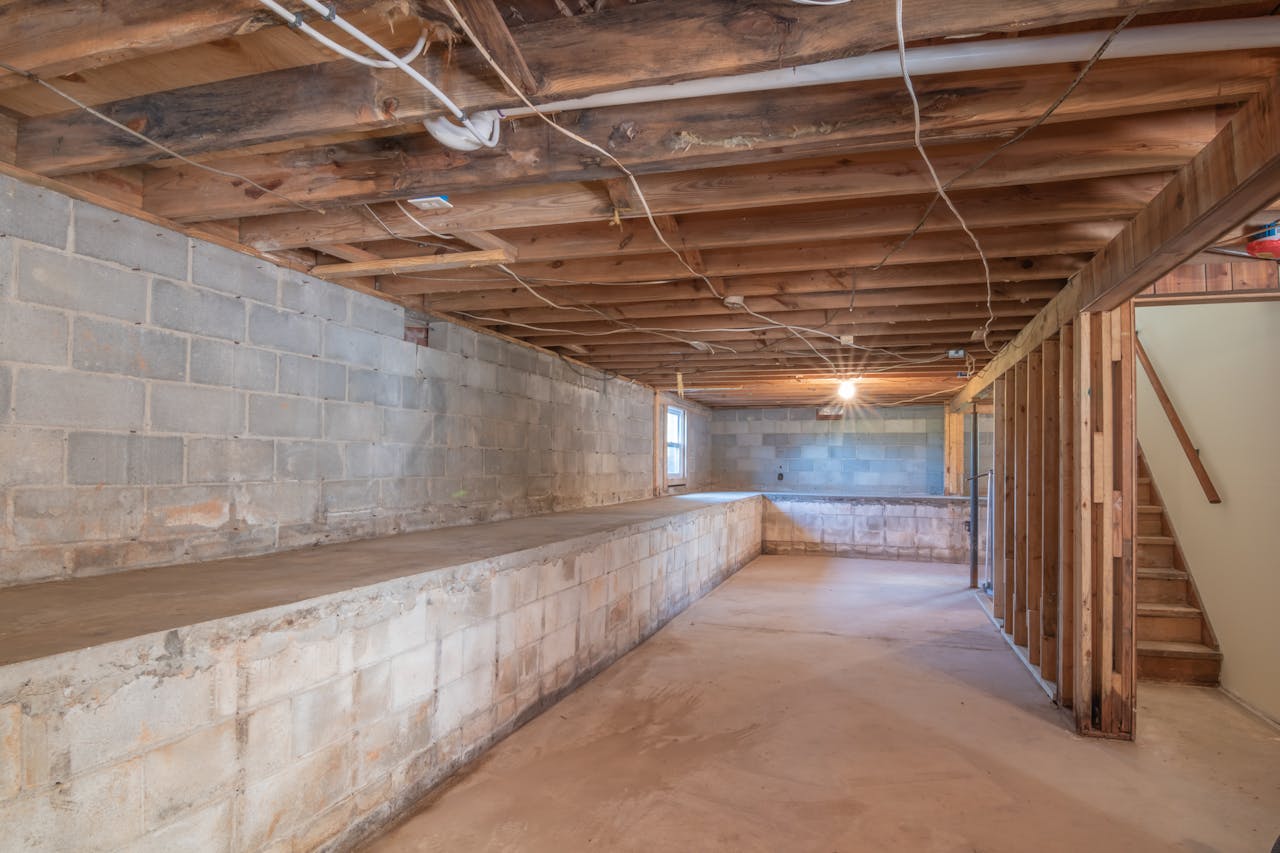 Unfinished basement with exposed wooden ceiling beams, bare concrete block walls, concrete ledge along one side, carpeted floor, open layout, wooden staircase, dim lighting