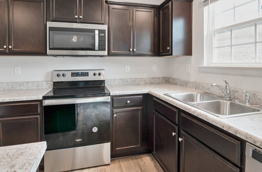 Modern kitchen interior, dark wooden cabinets, stainless steel appliances, electric stove with oven, mounted microwave, white tile backsplash, granite countertops, stainless steel sink, large window with natural light