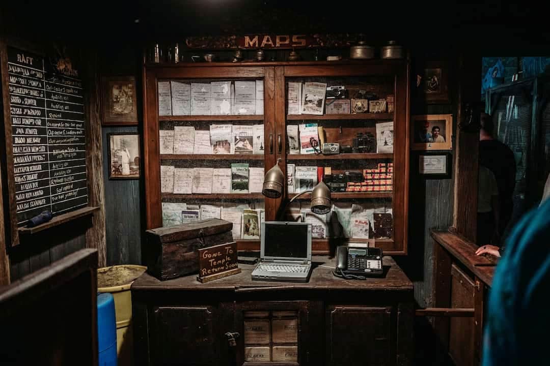 dimly lit vintage office, wooden cabinet labeled “MAPS”, old documents and film cameras on display shelves, antique desk with a closed laptop, telephone, desk lamps, wooden box labeled “Gone to temple return soon”, chalkboard list of raft services on the wall, framed photographs and certificates, rustic wooden walls and furniture