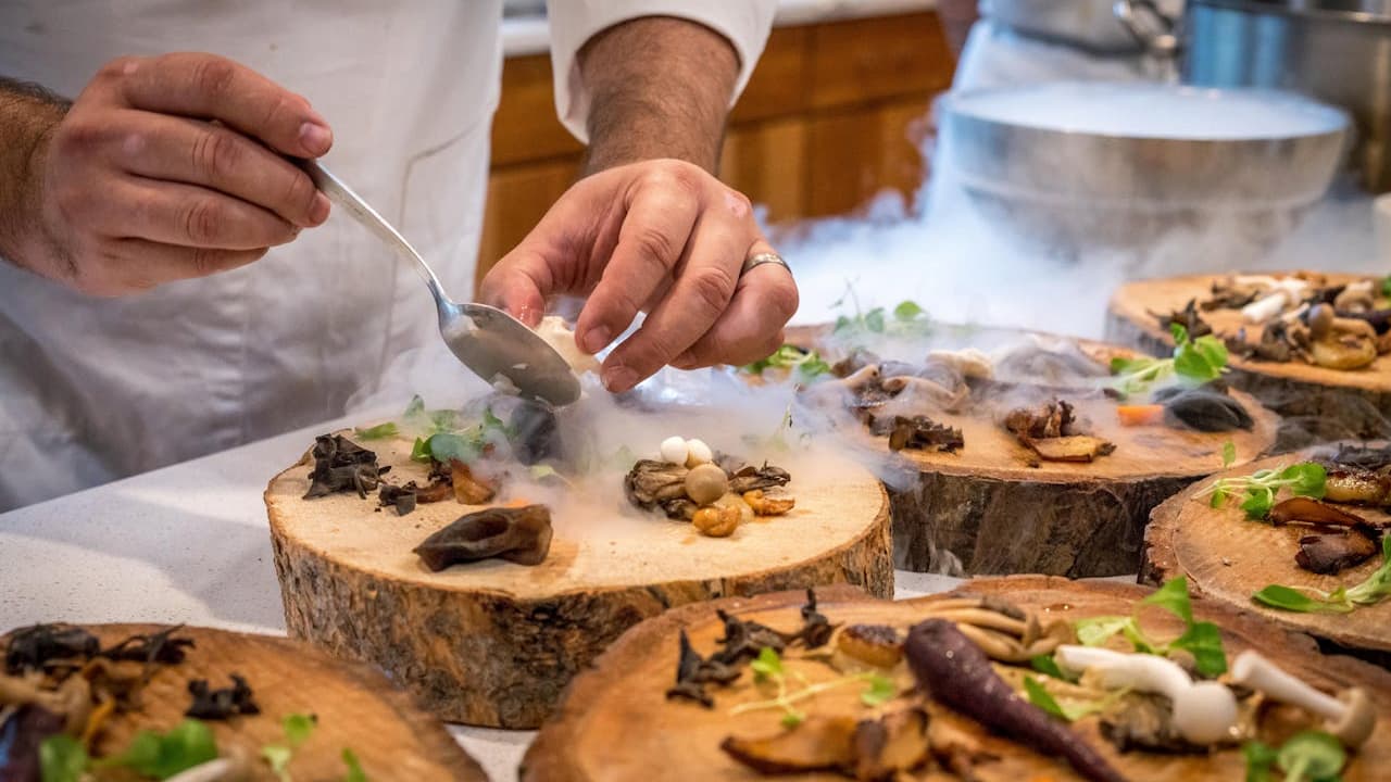 Chef preparing gourmet dishes, food presented on wooden slabs, wisps of smoke surrounding the plates, use of smoke infuser technique, fine dining setting, precise plating with mushrooms and microgreens