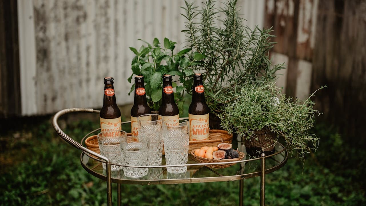 bar cart setup in a garden, wooden tray base, several bottles of dark glass drinks, two clear drinking glasses, surrounded by fresh green plants, rustic wooden background, soft outdoor lighting