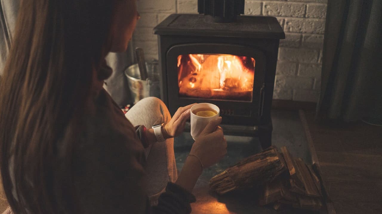 person holding mug, sitting near wood-burning stove, warm fire glowing, cozy indoor setting, dim lighting, relaxed atmosphere