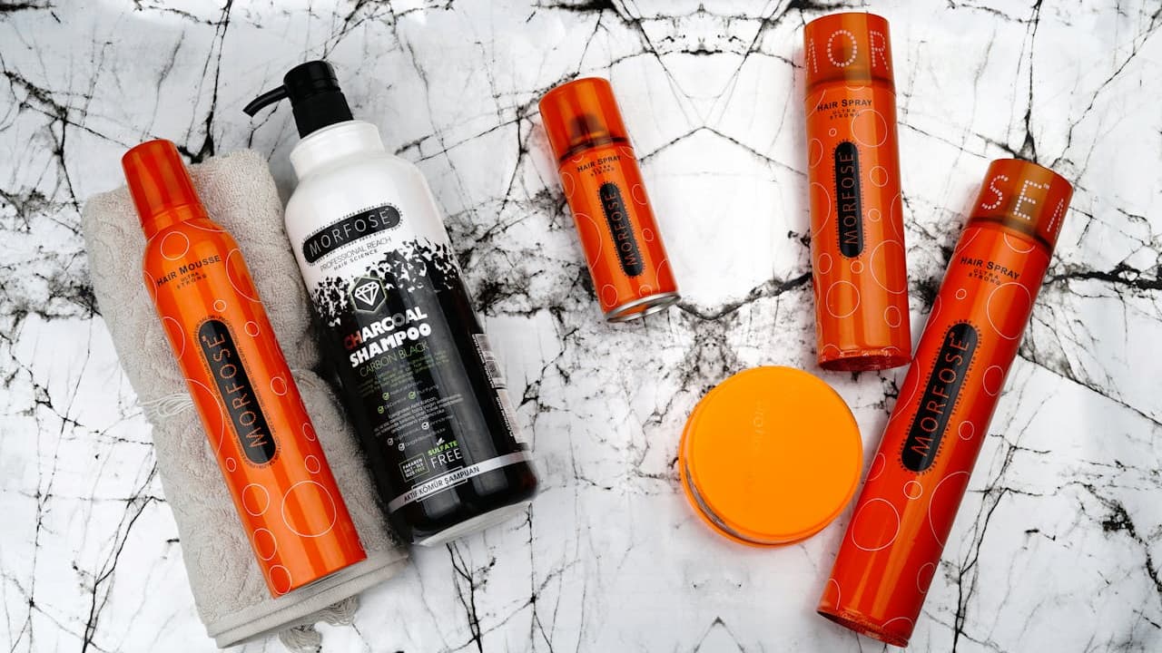 Assorted hair care products on a white marble surface, mostly in orange and black bottles, including spray cans, pump bottle, and a round container, arranged in a flat lay style