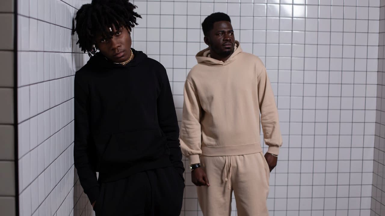Two men standing against a white tiled wall, one wearing a black hoodie and sweatpants, the other in a beige hoodie and sweatpants, both with hands in pockets, casual streetwear style