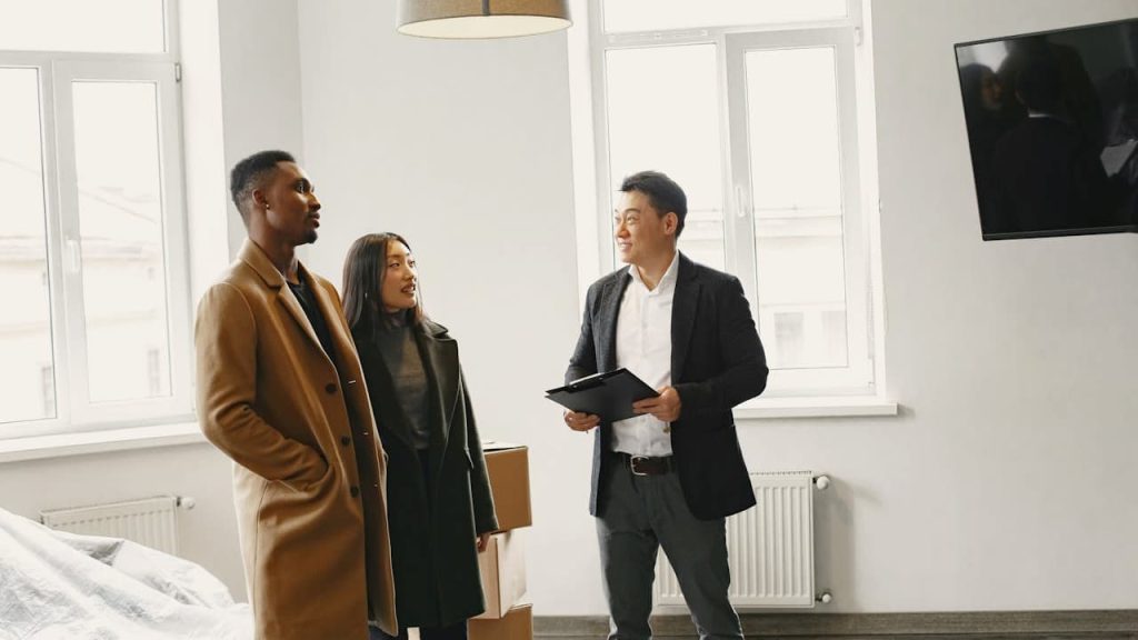 Real estate agent showing a modern, bright apartment to a couple, large windows letting in natural light, white walls, mounted flat-screen TV, minimal interior decor