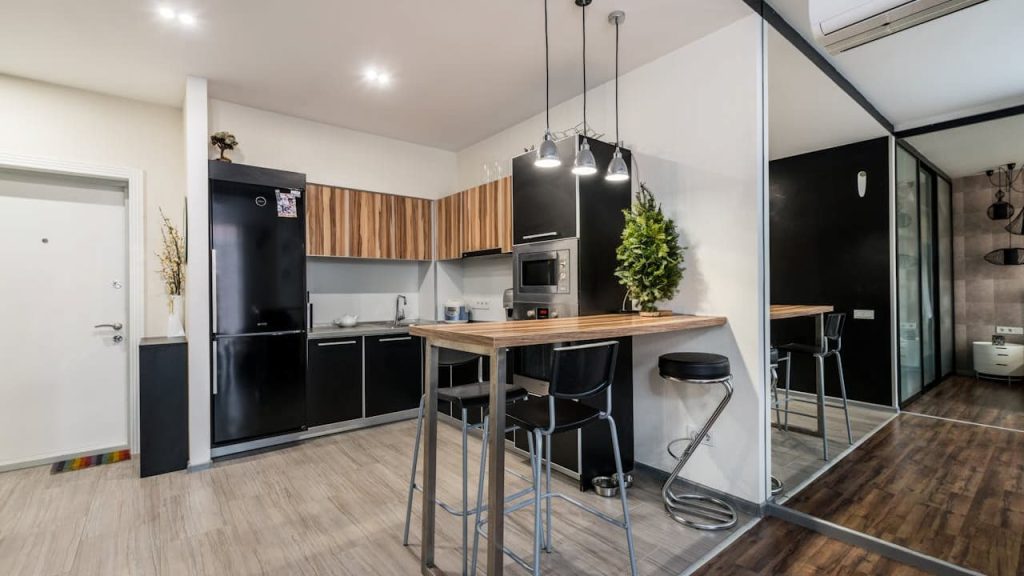 Modern kitchen interior, black appliances, wooden upper cabinets, light-colored countertop, bar-height dining table with metal legs, pendant lighting, potted plant for decor, tiled floor, white and black color scheme, open layout design