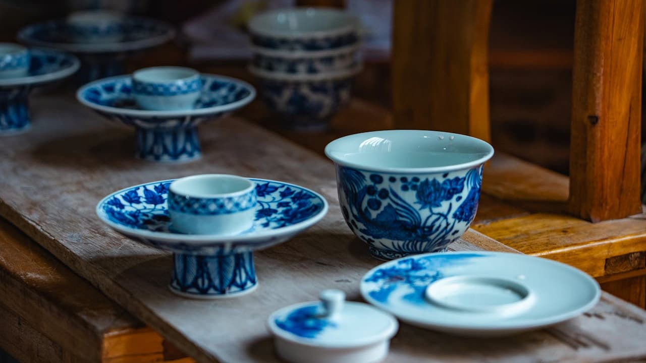 Assorted blue and white porcelain dishes on a wooden surface, including pedestal bowls, plates, and teacups, traditional floral and abstract patterns, warm indoor lighting, wooden shelves in the background