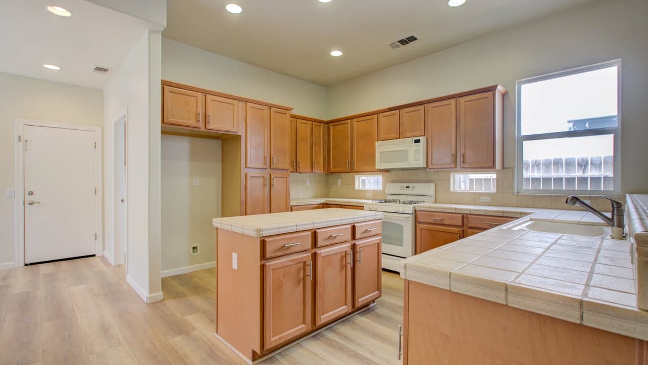 modern kitchen interior, wooden cabinets, tiled countertops, built-in appliances, overhead lighting, central island, light wood flooring, large window, neutral color scheme, open and clean layout