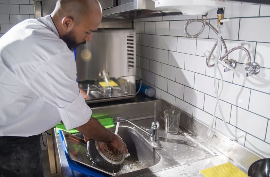 man washing a cast iron pan at a kitchen sink, wearing a white shirt and apron, tiled wall in the background, using a sponge under running water, surrounded by dish soap and utensils