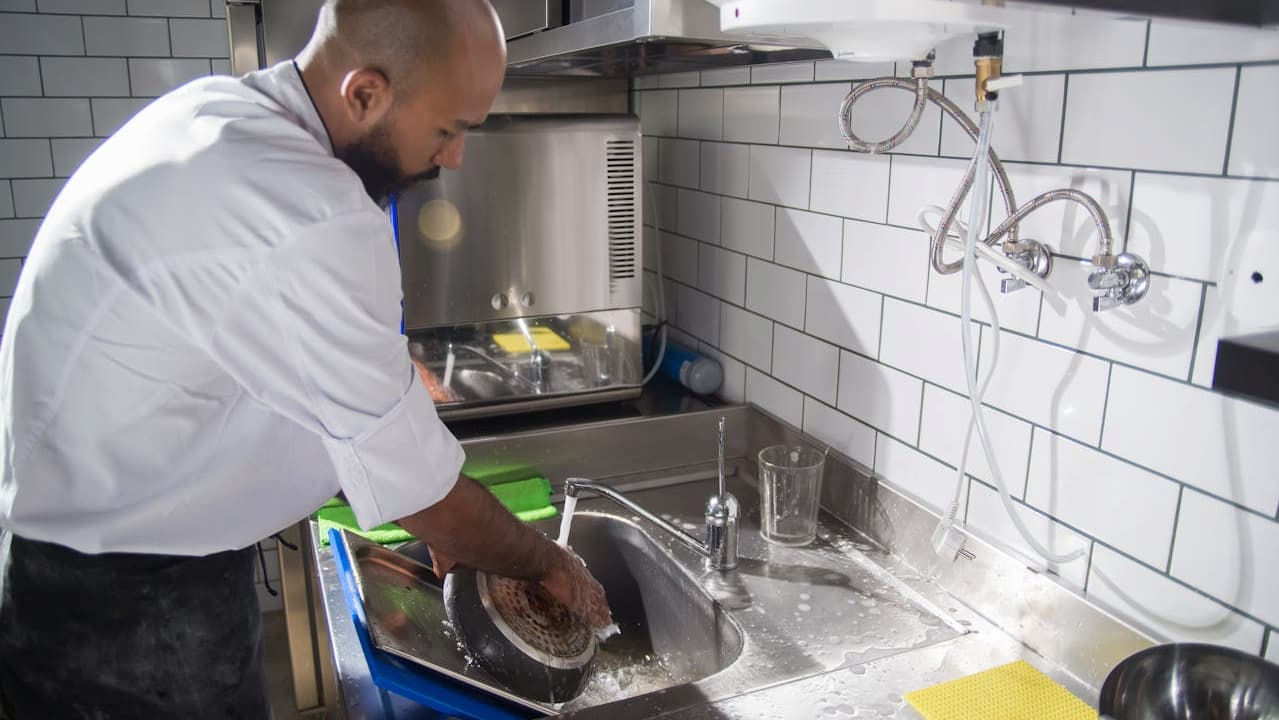 man washing a cast iron pan at a kitchen sink, wearing a white shirt and apron, tiled wall in the background, using a sponge under running water, surrounded by dish soap and utensils