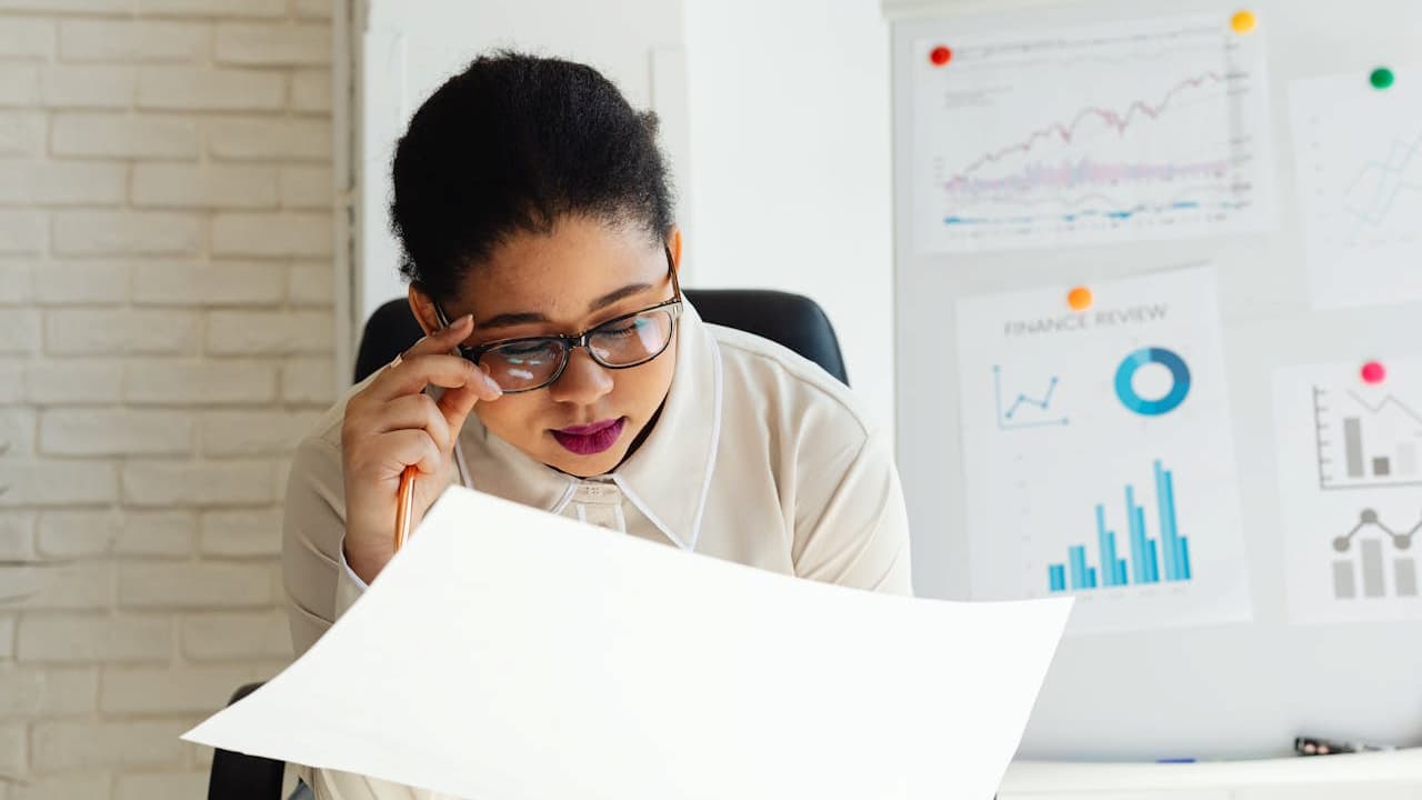 woman wearing glasses analyzing a large document, sitting at a desk, financial charts and graphs pinned on the board behind her, white and bright office setting, focused expression
