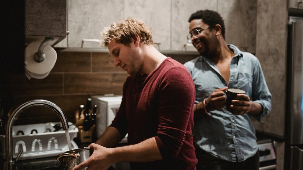 Two men in a modern kitchen, one washing dishes at the sink, the other holding a mug and smiling, casual clothing, relaxed atmosphere, warm indoor lighting, wooden backsplash, stainless steel appliances
