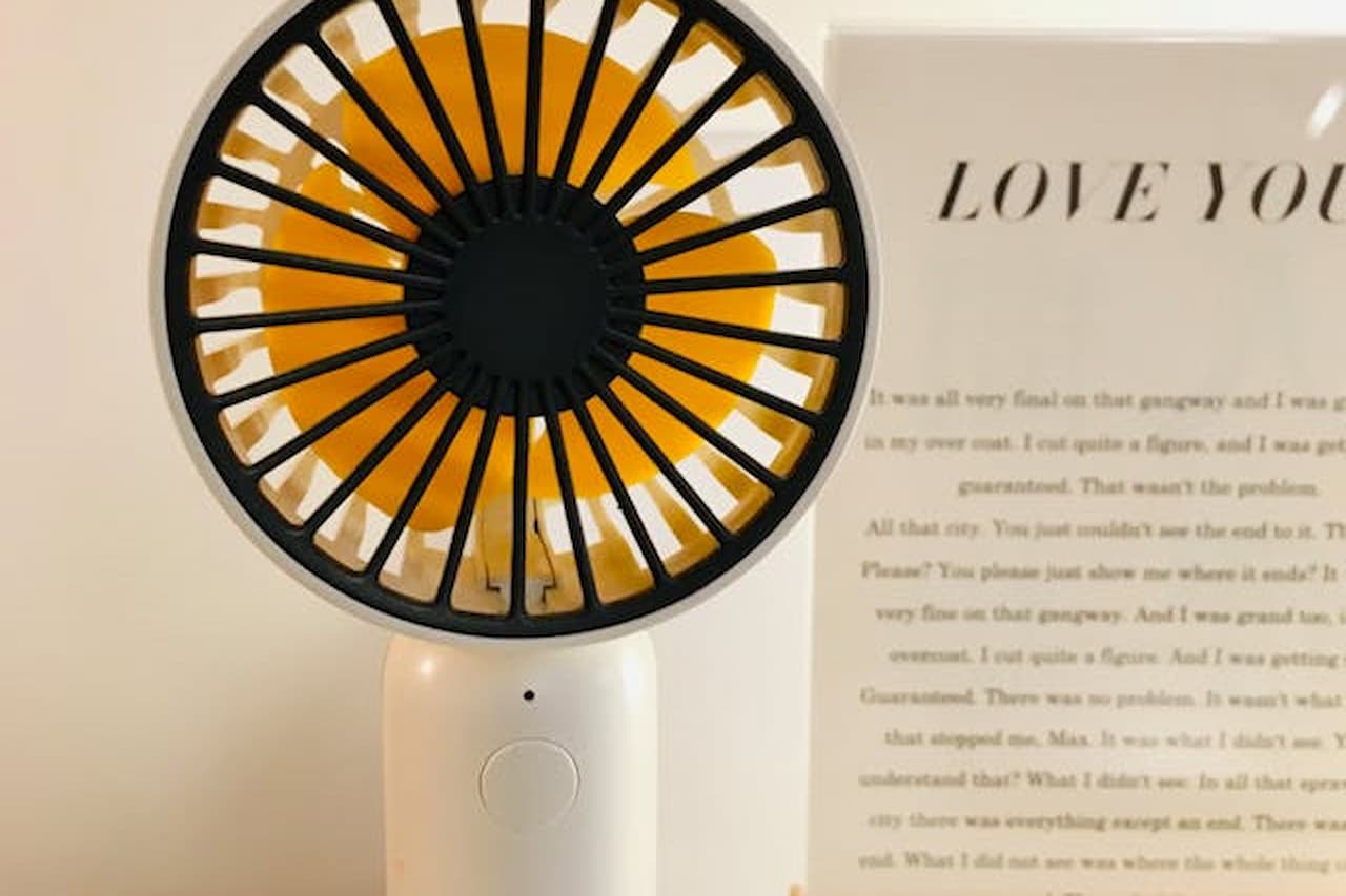 small portable fan, black and orange circular blades, white base, placed next to a printed text page, minimalistic indoor setting