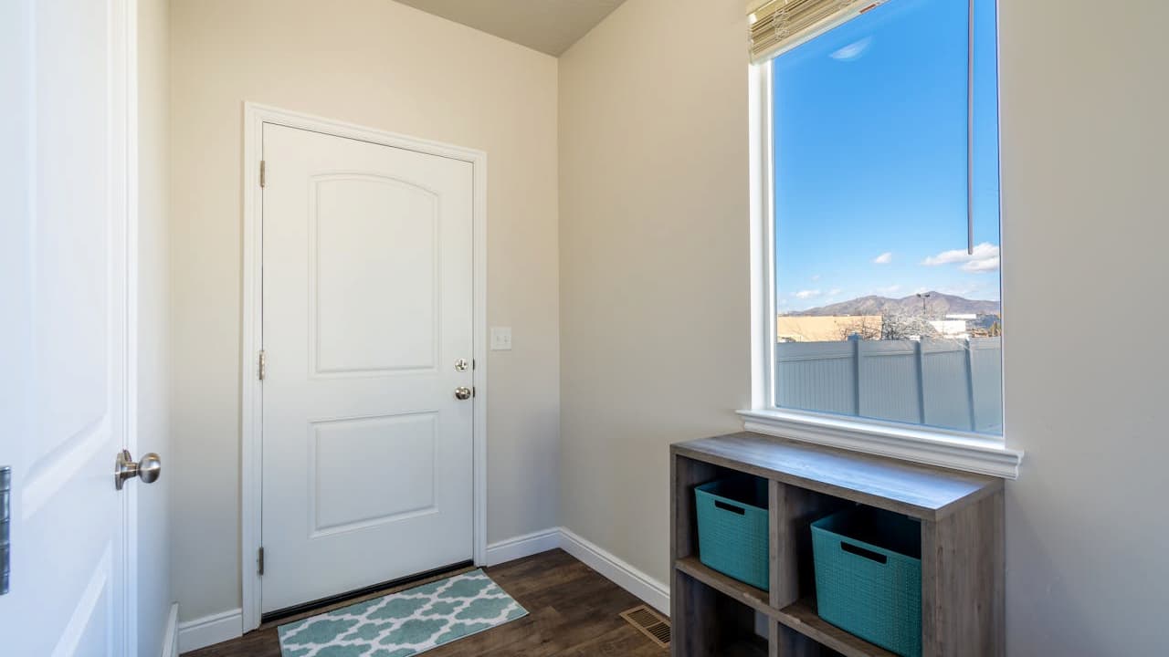 Small entryway with white walls, closed white door, wooden floor with a patterned rug, shelving unit with storage bins below a large window, blue sky and fence visible outside.