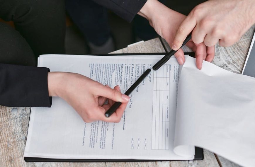 Two people reviewing a document, one hand holding a pen and pointing at the paper, the other flipping a page, visible text on the document, laptop nearby on a textured surface, collaborative work setting