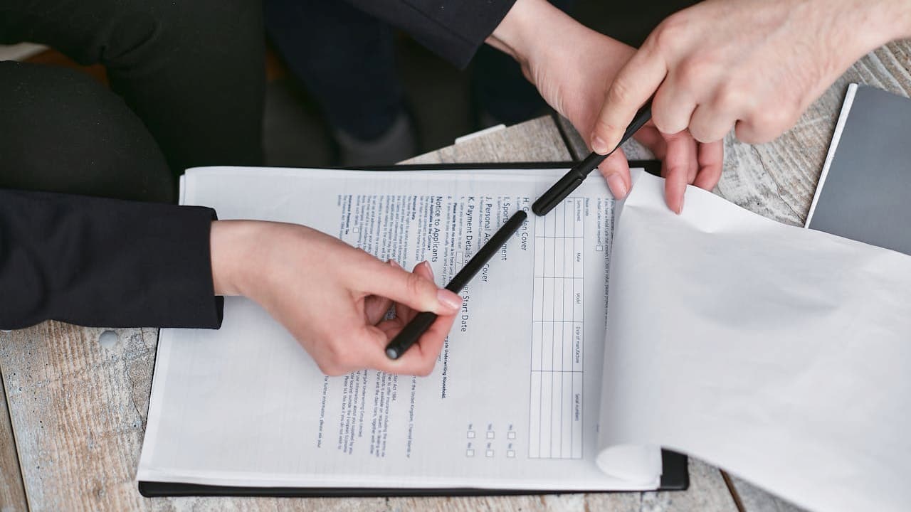 Two people reviewing a document, one hand holding a pen and pointing at the paper, the other flipping a page, visible text on the document, laptop nearby on a textured surface, collaborative work setting