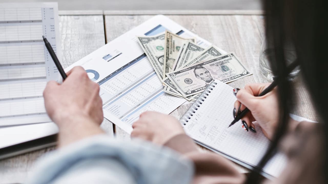 two people reviewing a printed map with notes and markings, one person pointing at the map, the other holding a pen, close-up view, document partially visible on the side, wooden table surface underneath