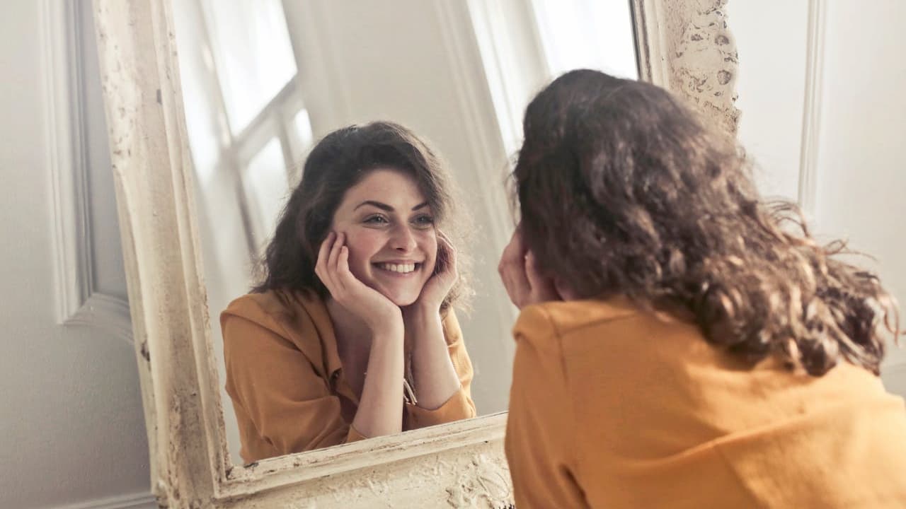 Woman with curly brown hair, wearing a mustard yellow top, smiling at her reflection, leaning on a vintage-style mirror, soft natural light coming through window blinds in the background