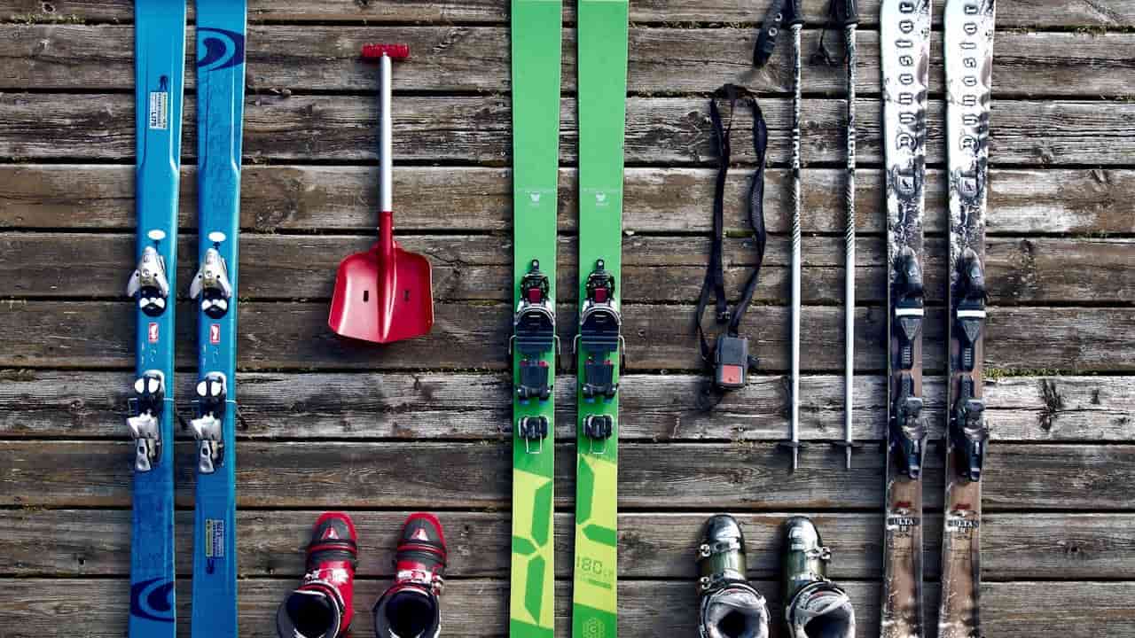 assorted ski equipment, three pairs of skis, ski poles, ski boots, red snow shovel, wooden deck background, gear laid out in rows, winter sports gear