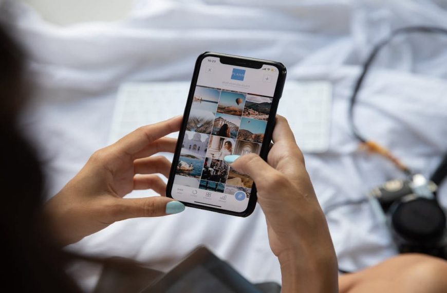 Person holding smartphone, scrolling through social media feed, visible travel or lifestyle posts on screen, phone held over a white bedsheet, keys and headphones beside the phone, casual indoor setting, suggesting travel or relaxation