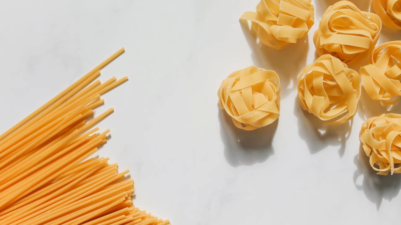 Uncooked pasta varieties on a white surface, long ridged spaghetti on the left, round nest-shaped pasta on the right, dry, golden yellow in color