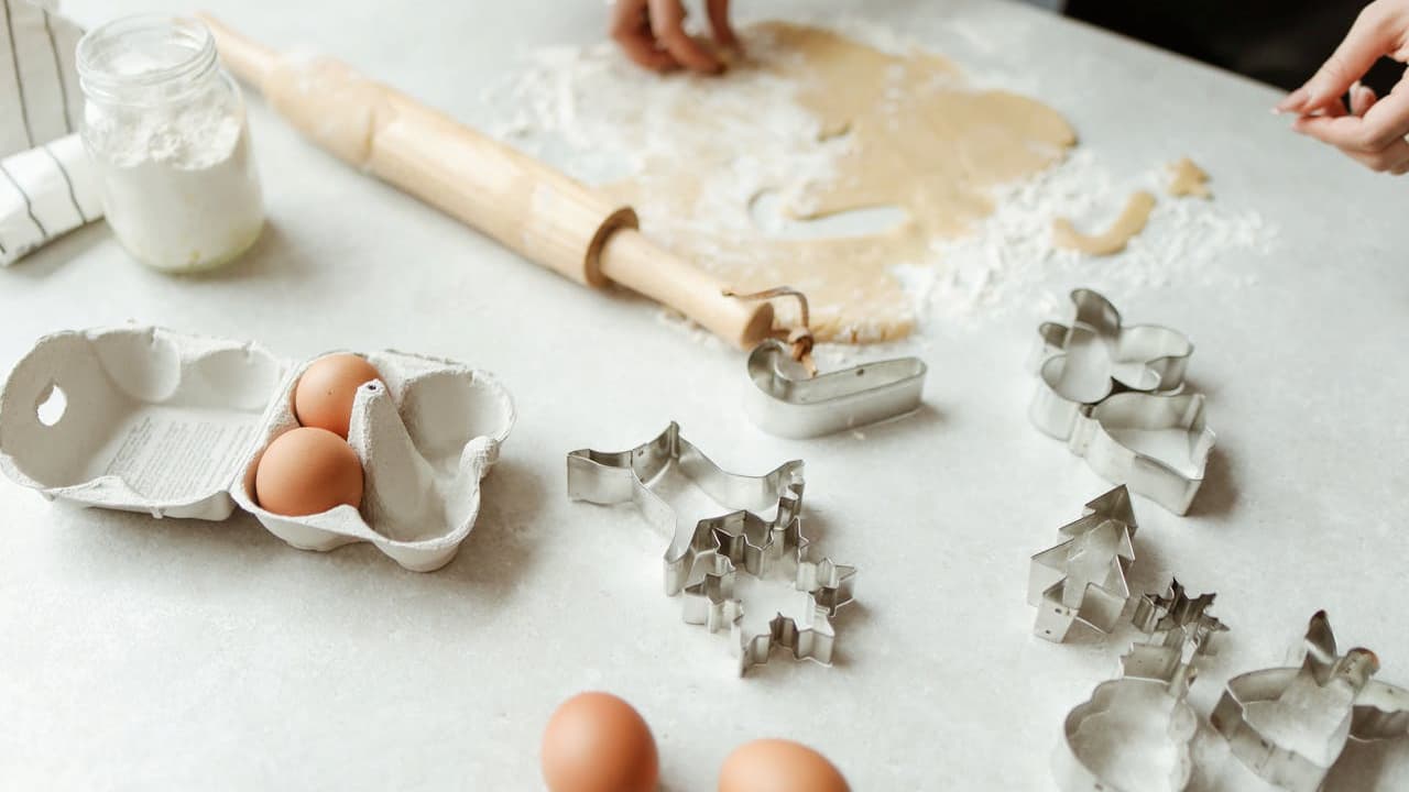 Hands preparing dough, rolling pin on floured surface, cracked eggs and shells, cookie cutters in various shapes, baking in progress, white countertop, homemade pastry preparation