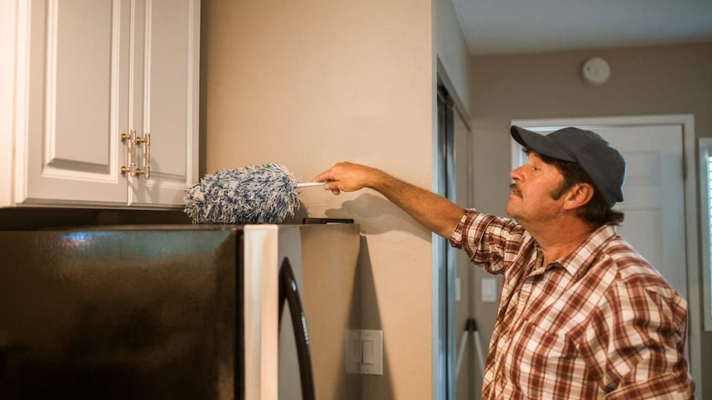 man removing cobwebs, long-handled duster, reaching ceiling corner, casual clothing, indoor cleaning, high wall area, bright interior