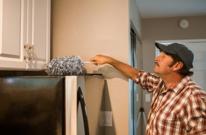 man removing cobwebs, long-handled duster, reaching ceiling corner, casual clothing, indoor cleaning, high wall area, bright interior