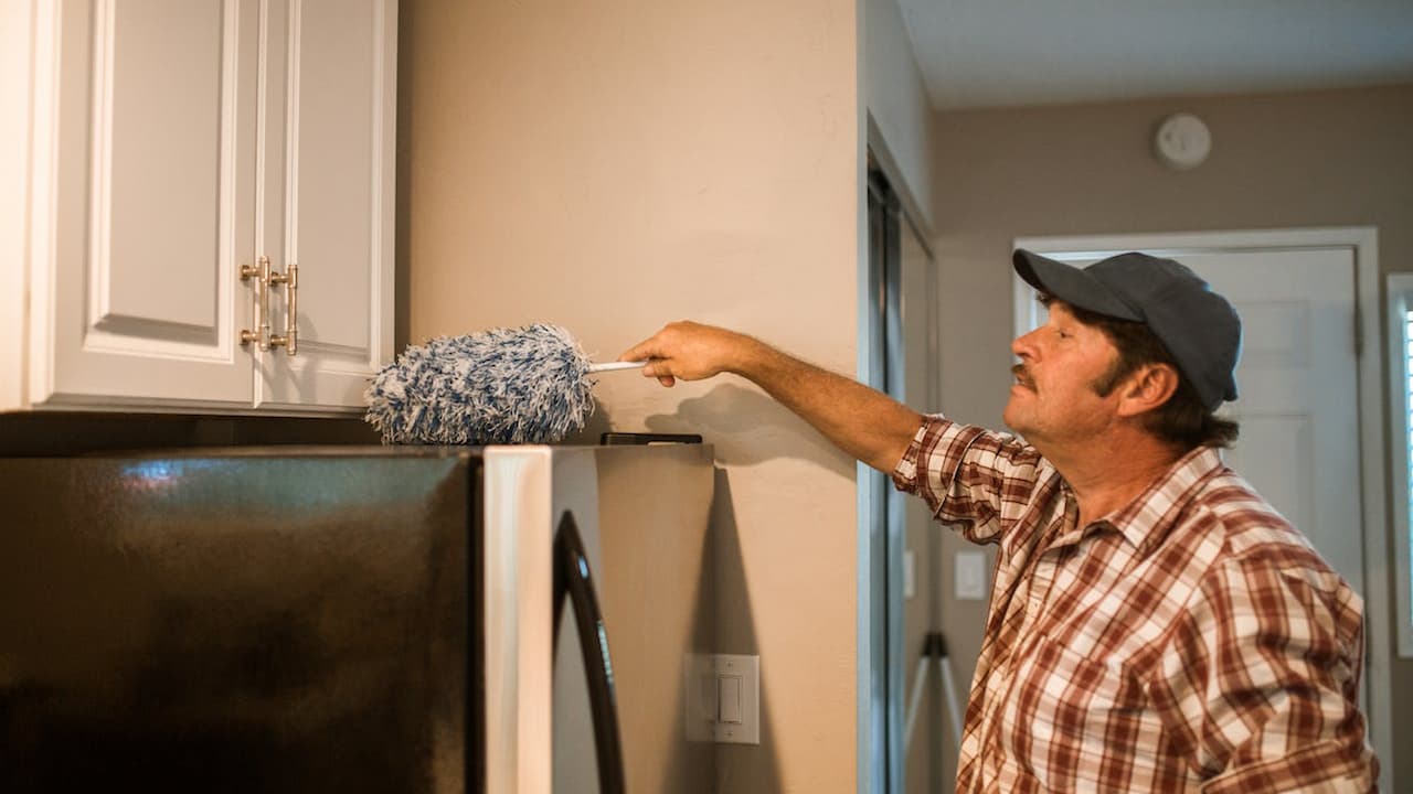 man removing cobwebs, long-handled duster, reaching ceiling corner, casual clothing, indoor cleaning, high wall area, bright interior