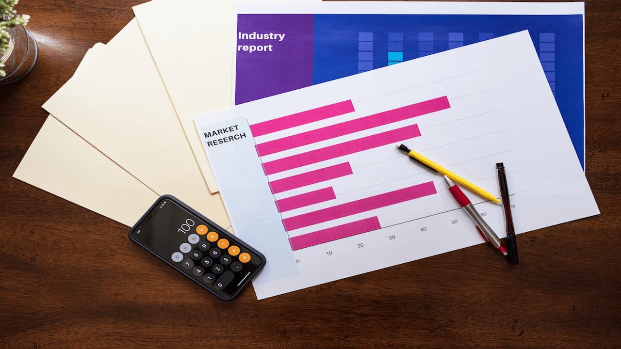 Printed charts on a desk, pink bar graph on white paper, manila folders underneath, digital calculator nearby, two pens placed diagonally, and a colorful chart visible in the background