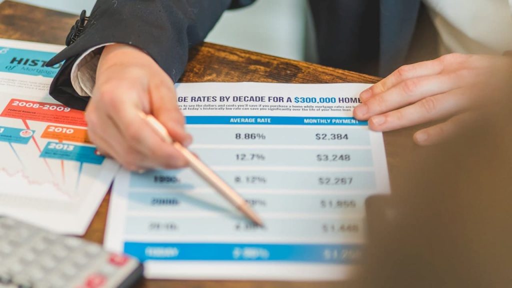 person holding a financial document showing average mortgage rates and monthly payments for a $300,000 home, pointing at data with a pen, another hand holding the paper, historical mortgage rate chart visible on the table, calculator partially in view