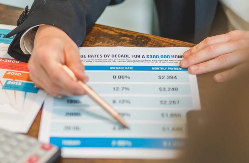 person holding a financial document showing average mortgage rates and monthly payments for a $300,000 home, pointing at data with a pen, another hand holding the paper, historical mortgage rate chart visible on the table, calculator partially in view