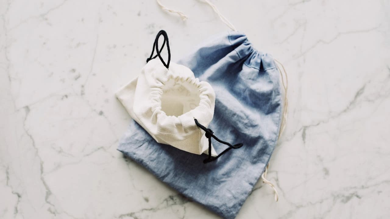 Reusable cloth face mask with ear loops, placed on a folded light blue knitted beanie, white marble surface background, minimalist flat lay style